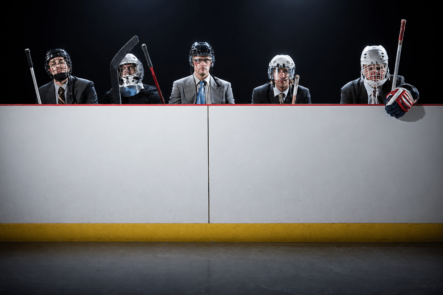 Men dressed in suits and hockey masks, holding hockey sticks, lined up behind ice-rink barrier