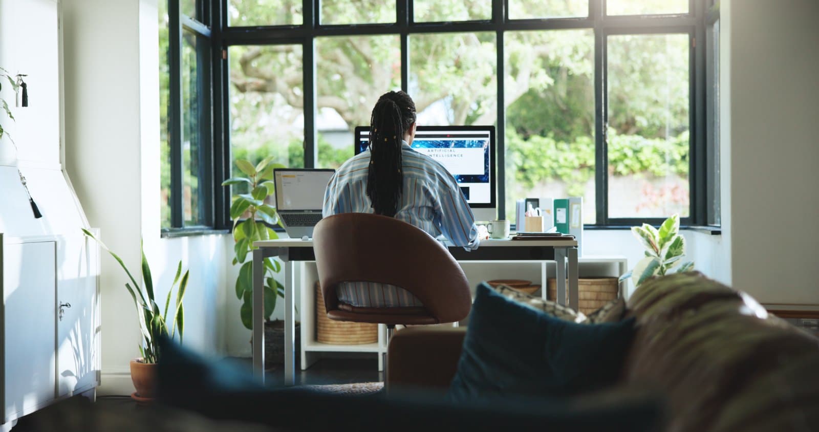 A person working on a computer from their home, they are sat in front of a window with a view into a garden.